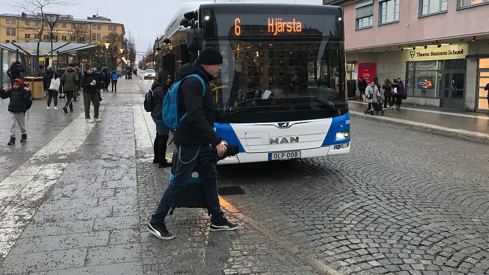 En kollektivtrafikbuss, nummer 6 mot Hjärsta, står parkerad i mitten av bilden i stadsmiljö, framför bussen passerar en person.