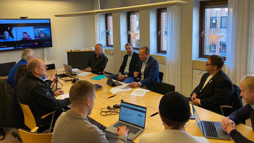 Politiker håller presskonferens vid ett stort avlångt bord. Fyra-fem politiker på ena långsidan. Fyra-fem journalister på den andra sidan.