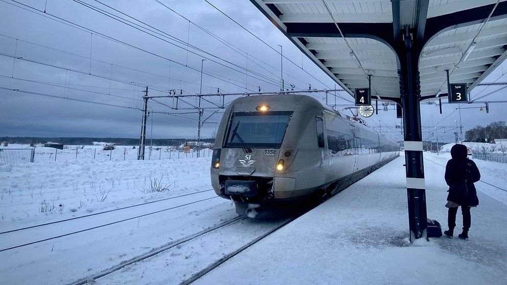Ett modernt tåg anländer till en snöig perrong med en ensam passagerare stående bredvid spår 3. Himlen är mulen och omgivningen täcks av ett vinterlandskap.