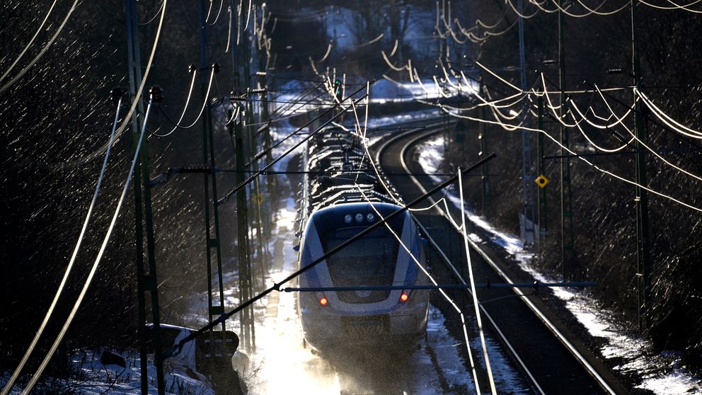 Ett persontåg åker längs en kurviga järnvägslinje i ett vintrigt landskap, omgiven av snötäckta träd och elektriska stolpar. Dämpad belysning skapar en mystisk atmosfär.