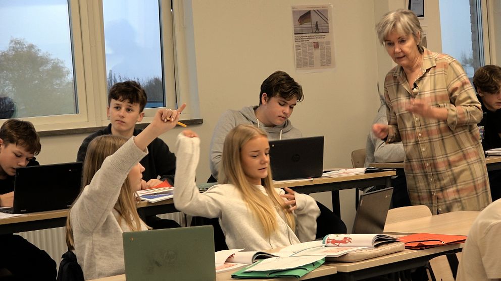 En klassrumssituation där en lärare förklarar något för eleverna. Två elever i förgrunden, en av dem räcker upp handen för att ställa en fråga. Flera elever sitter vid sina skrivbord med bärbara datorer framför sig. Fönster i bakgrunden släpper in naturligt ljus.