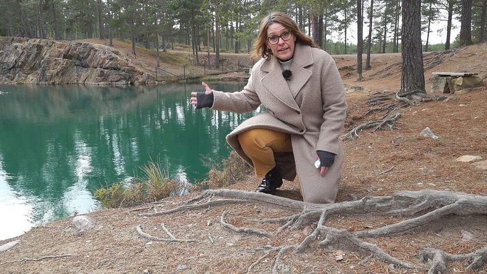 Kvinna i beige kappa och brandgula byxor står på knä framför en grön sjö i en skog