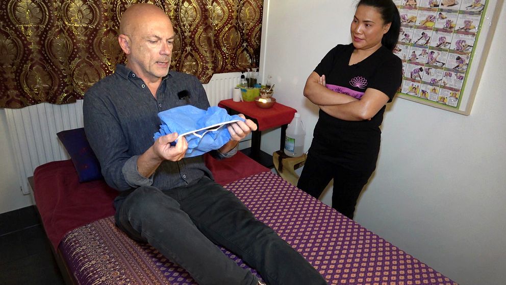 En man sitter på en massagebänk och håller en blå handduk i sina händer. I bakgrunden står en kvinna med armarna i kors. Rummet är inrett med en mönstrad kudde och en dekoration på ett bord, samt en vägg prydd med bilder.