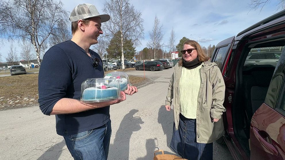 En kvinna står vid en bil och en man står och håller i två tårtor. Personerna ler mot varandra.