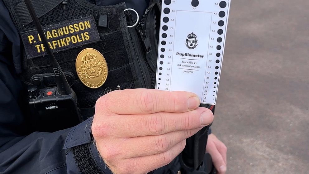 En trafikpolis håller upp en pupillometer, ett verktyg för att mäta pupilens storlek, med text som visar att det används av Rikspolisstyrelsen. Polisen är klädd i uniform med en namnskylt som tydligt anger att personen är P. Magnusson.