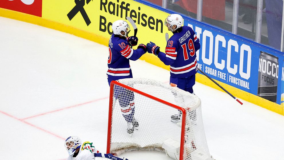 Två hockeyspelare firar mål bakom målbur, målvakt sitter på isen framför dem.