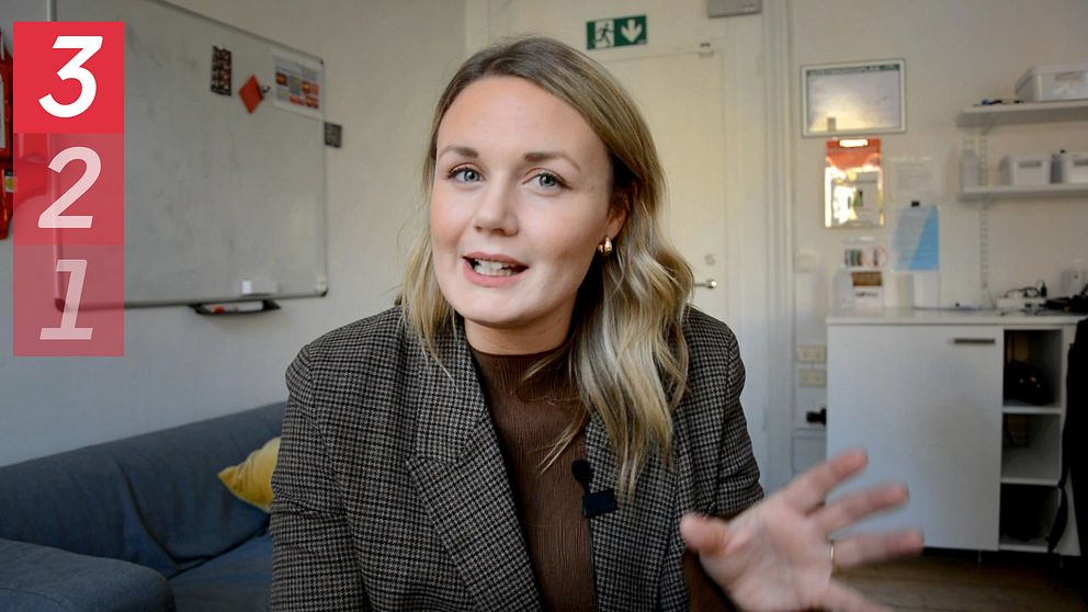 En person sitter i en modern inomhusmiljö, iförd en rutigt kavaj, och gestikulerar med en hand. I övre vänstra hörnet finns en grafik med siffrorna 3, 2, 1 i rött.