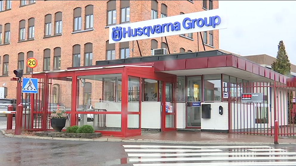 Entrén till Husqvarna Groups anläggning, med en röd entréport och skyltar som visar företagets namn. I bakgrunden syns en tegelbyggnad och trafikskyltar. Det är en regnig dag med våta markytor.