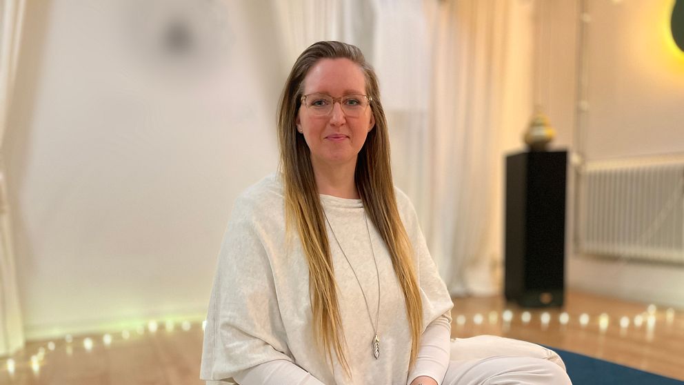 En person sitter i en meditationsställning på en blå matta i ett rum med dämpad belysning. Personen har på sig vita kläder och en lång halskedja. Bakgrunden visar ljusa gardiner, en upplyst växt och ett element.
