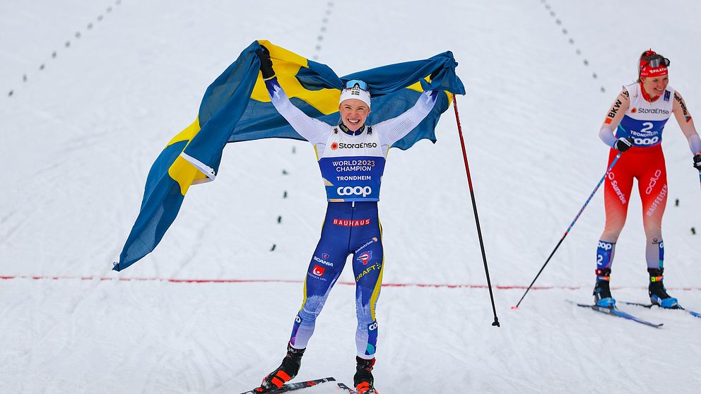 Svensk längdskidåkare firar med flagga, vid mållinjen under tävling.