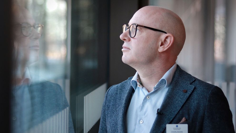 En person i en grå blazer står vid ett fönster och ser ut mot utsidan, med reflektioner synliga på glaset. Personen har en namnskylt fäst på blazern. Bakgrunden visar suddiga former av det som kan vara en modern miljö.
