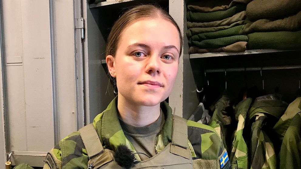 En person i militäruniform står i en klädkammare med grön och svart kamouflage. I bakgrunden syns flera militärplagg som hänger prydligt. Personen bär en mikrofon och har en Sveriges flagga på sin uniform, vilket indikerar att de tillhör den svenska militären.