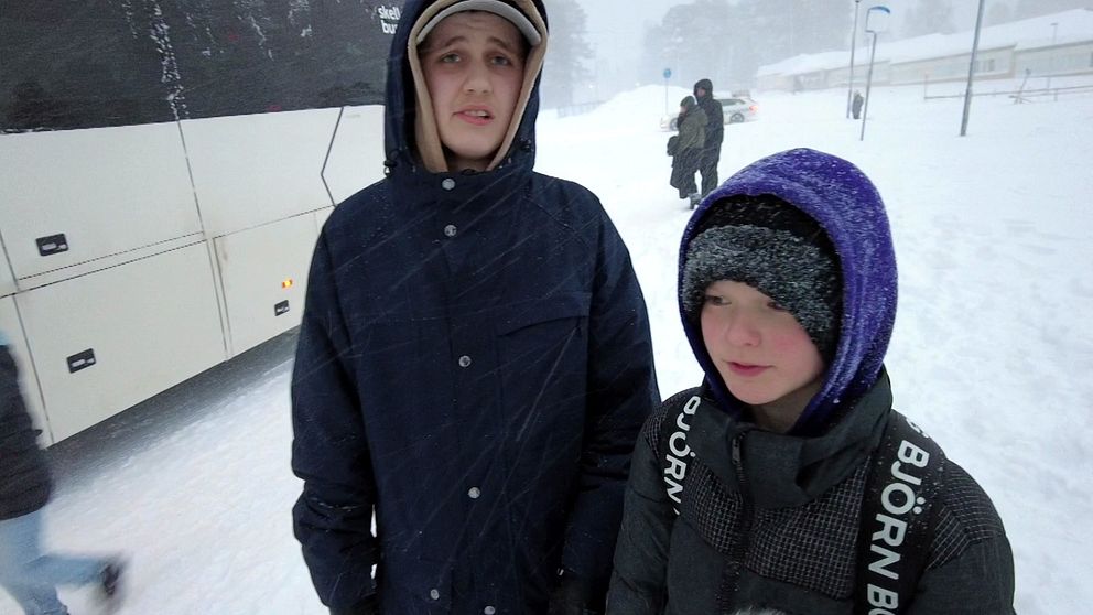 Skolbarnen Alvar och Liam  står framför en vit buss med huvorna uppdragna. Det snöar kraftigt.