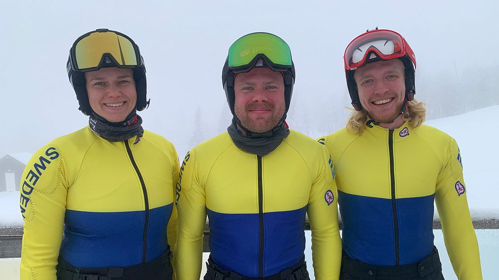 Tre personer i gul- och blåklädda tävlingsdräkter, representerande Sverige, står bredvid varandra i en dimmig utomhusmiljö. De bär hjälmar och handskar som tyder på att de deltar i en vintersport.