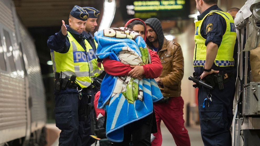 Poliser interagerar med en person som håller ett barn insvept i en filt på en tågstation, medan en andra polis övervakar situationen. I bakgrunden står ett tåg stilla på perrongen.