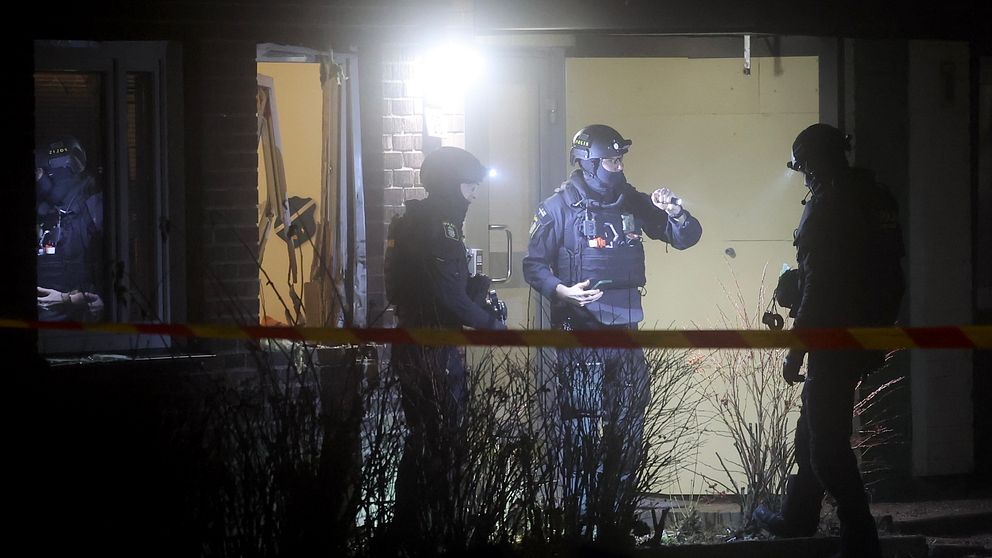 Poliser iklädda taktisk utrustning står utanför ett upplyst rum, med avspärrningsband i förgrunden, medan de genomför en insats på natten.