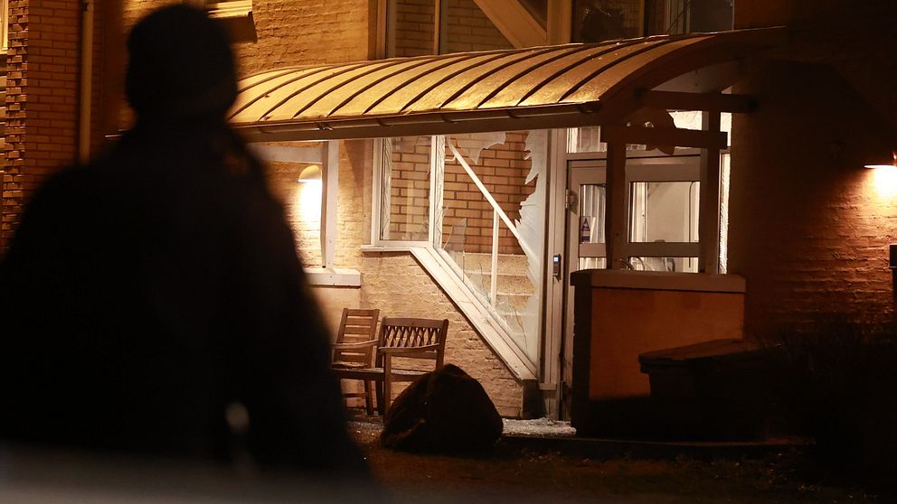 En suddig siluett av en person ses i förgrunden med ett belyst flerfamiljshus i bakgrunden med krossade rutor. Ett avspärrningsband syns nära marken framför personen.