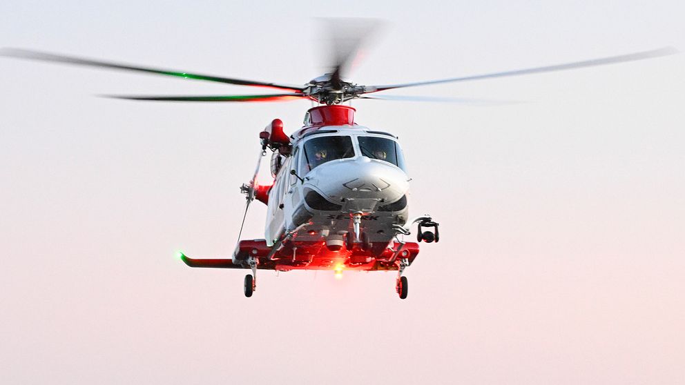 En räddningshelikopter flyger i luften under skymning med en person synlig vid sidan av helikoptern.