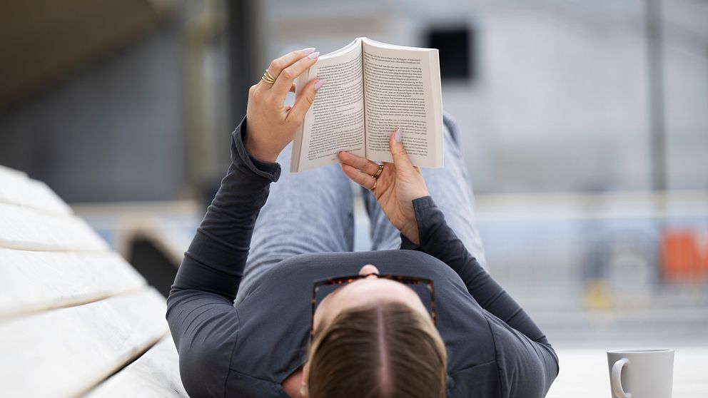 Person ligger på rygg utomhus och läser en bok, kaffemugg står bredvid.