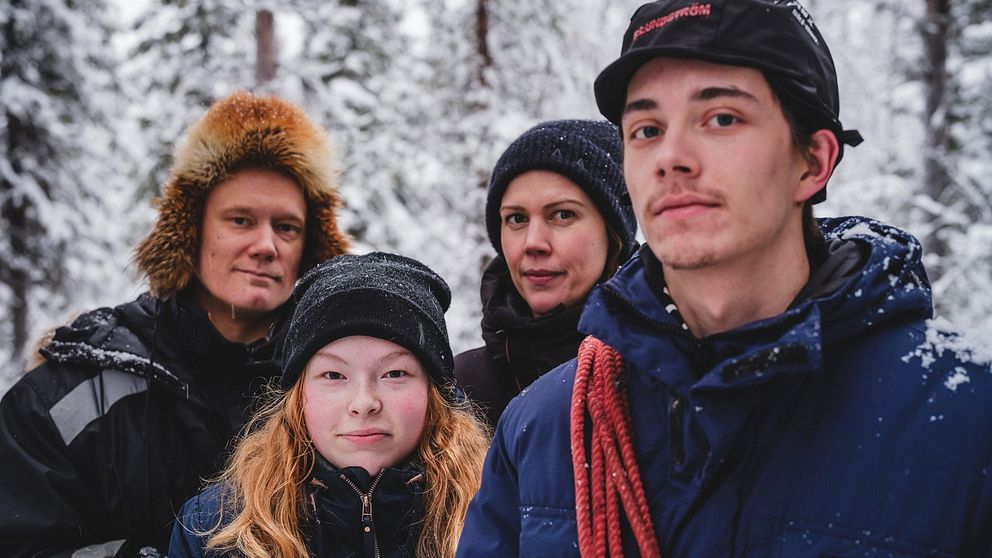 Fyra personer som står utomhus i ett snöigt skogslandskap, klädda i varma vinterkläder. Personen längst till höger bär en mörk blå jacka med en röd rep över axeln och en mörk mössa med skärm. Träden i bakgrunden är täckta av snö.