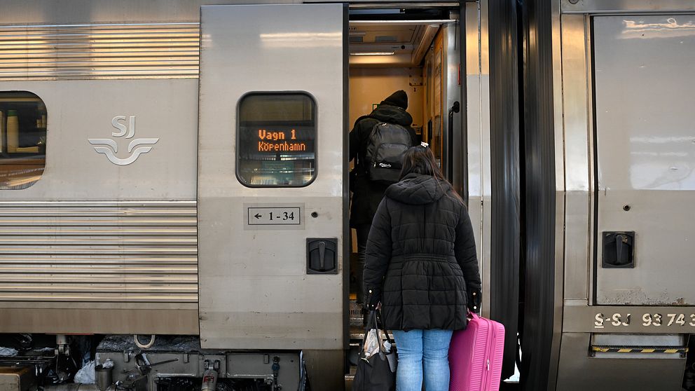 En resenär med en rosa resväska går ombord på ett tåg vid en plattform, med tågdörren öppen och en annan passagerare som redan har klivit in. Tåget är märkt med SJ och destinationen Köpenhamn.