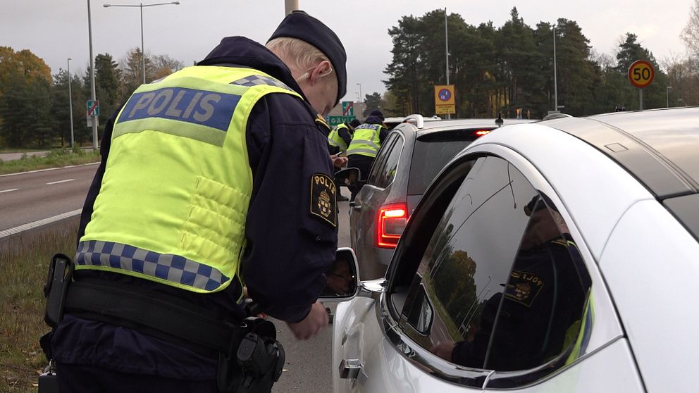 En polis i uniform genomför en trafikkontroll och pratar med en bilförare genom fönstret på en rad av bilar som står stilla på en landsväg.