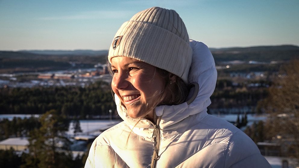 Längdskidåkaren Johanna Hagström iklädd en vit jacka och en ruta med en stickad mössa står vid en utsiktsplats i Falun. Bakgrunden visar en vinterlandskap med snö, träd och en fjärran vy av täckta kullar och en sjö. Dagsljuset skapar en klar och kall atmosfär.