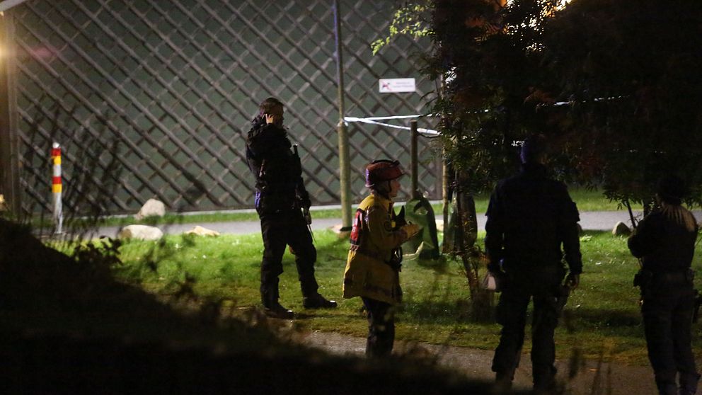 Flera personer i uniform står på en grön yta under natten. En person är klädd i en gul jacka medan de andra bär mörka kläder. De verkar vara i en beredskapssituation, med fokus på området bakom dem, där ett staket syns. Belysning från en lampa bidrar till den svaga synligheten.