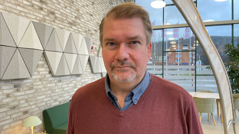 En person står inomhus framför en tegelvägg med geometriska dekorativa paneler, ett modernt mötesrum med en soffa och fönster som visar en stadsbild utanför.