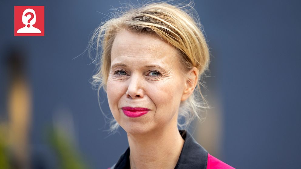 En kvinna med blont, halv långt hår bär en svart och rosa outfit. Bakgrunden är suddig och består av en grå vägg med gröna växter i närheten, vilket skapar en avslappnad atmosfär.