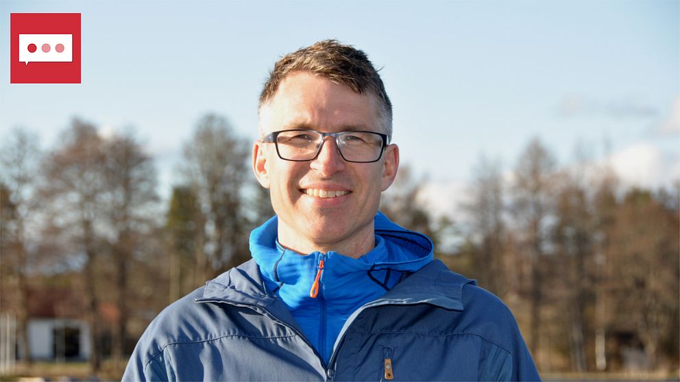 En person i en blå jacka står framför en sjö eller en stor vattensamling, med klart väder och träd i bakgrunden. Det ger en känsla av utomhusaktiviteter och natur.