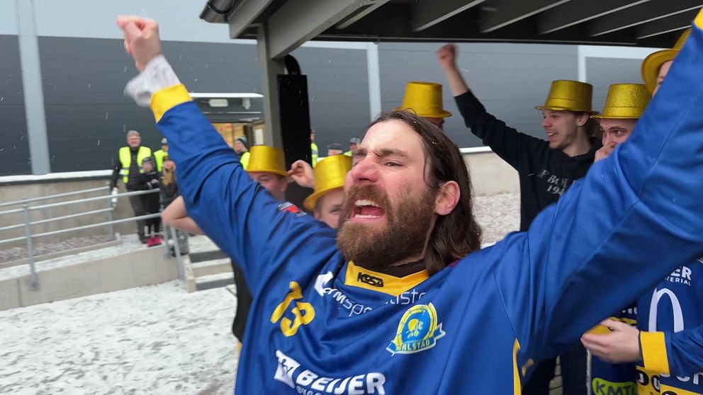 Flera personer firar utomhus, klädda i blå och gula sporttröjor nummer 13 och 10 samt guldhattar, med snö eller hagel på marken. I bakgrunden syns säkerhetspersonal och en byggnad.