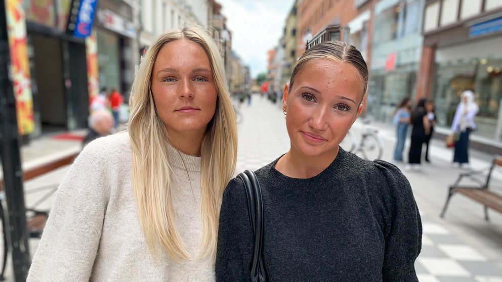 Två tjejer står på en shoppingata och tittar in i kameran.