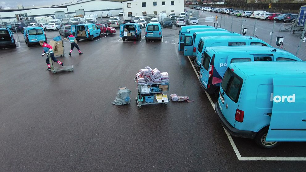En företagsparkering med flera blå eldrivna transportbilar, där några är öppna och lastade med paket. I förgrunden finns en lastpall med färgglada paket och försändelser, medan två arbetare i reflexvästar förbereder lastning. Området är vått, vilket tyder på nyligen fallande regn.