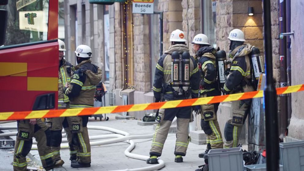 Fyra brandmän i skyddsutrustning står utanför en byggnad på Södermalm i Stockholm, omgivna av brandbilar och brandslangar. De diskuterar situationen medan en av dem håller en släckutrustning. En av brandbilarna syns till vänster, och ett avståndsband markerar avspärrningen runt området.