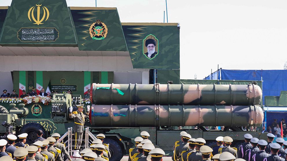 Militärparad i Iran, med soldater och lastbil som bär missiler framför byggnad.