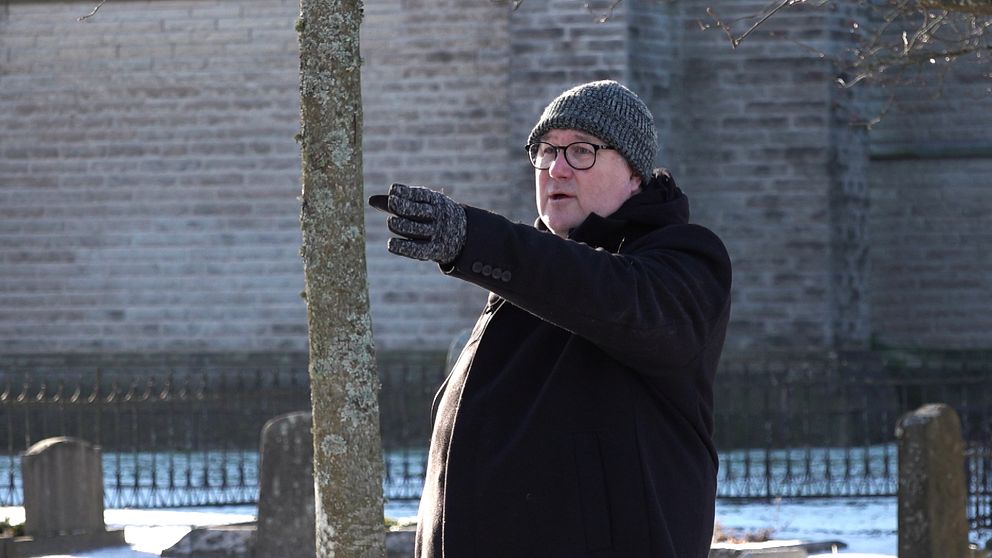 En person står utomhus, varmklädd i en mörk jacka och mössa, och pekar med handen. Bakgrunden består av en mur och ett träd, med snö på marken.