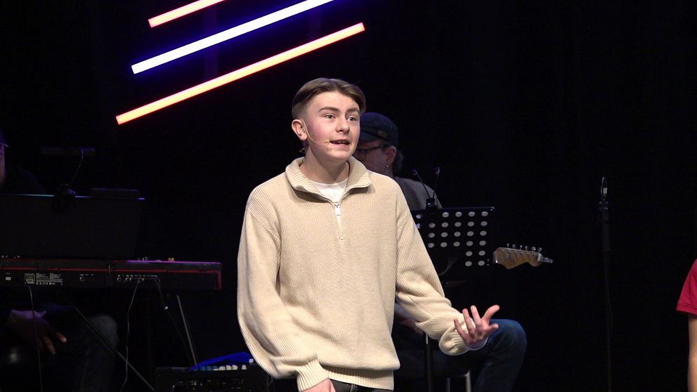 En ung man står på en scen  under en musikalisk framträdande. Bakom honom syns musiker som spelar instrument. Scenens belysning består av färgglada lysrör.
