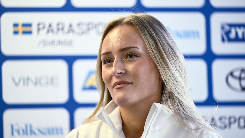 En person iklädd en vit jacka med ”Parasport Sverige” logotyp står framför en bakgrund med olika sponsorer inom idrott.