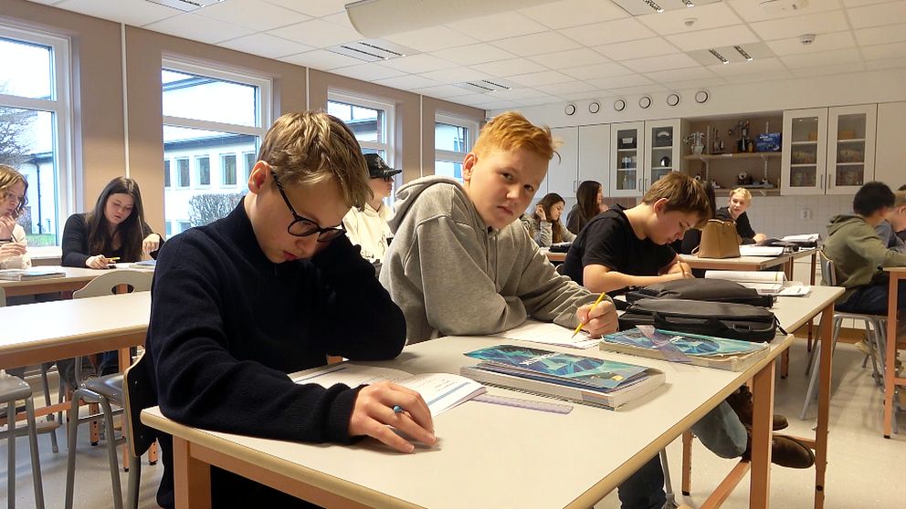 Elever sitter vid skrivbord i ett klassrum och arbetar med skoluppgifter. Tre elever i förgrunden är fokuserade på sitt arbete, med skolböcker och skrivhäften framför sig. Bakom dem finns fler elever, alla i ett ljust rum med stora fönster och klassrumsmöbler.