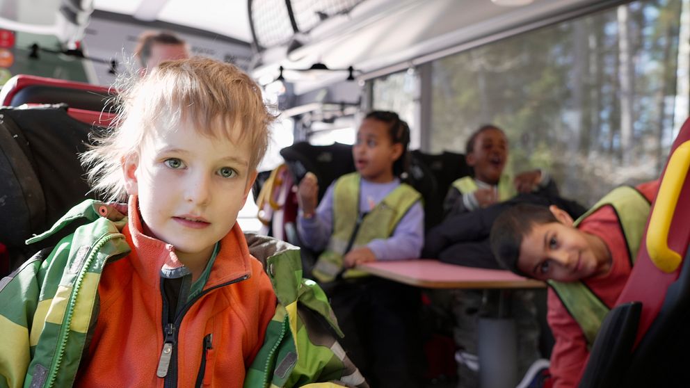Barn sitter i en buss med reflexvästar, soligt utomhus. Ett barns ansikte är suddat.