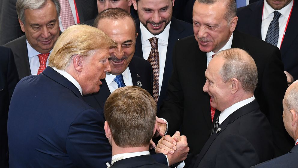 Donald Trump och Vladimir Putin skakar hand och deltar i en konversation bland en grupp män i kostymer.