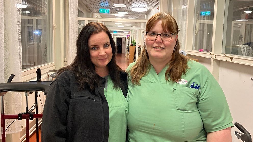 Saana Mänttäri och Anna-Lena Ramsten står i arbetskläder tillsammans framför en ljus korridor. I bakgrunden syns fönster och en rullstol i en vårdmiljö.