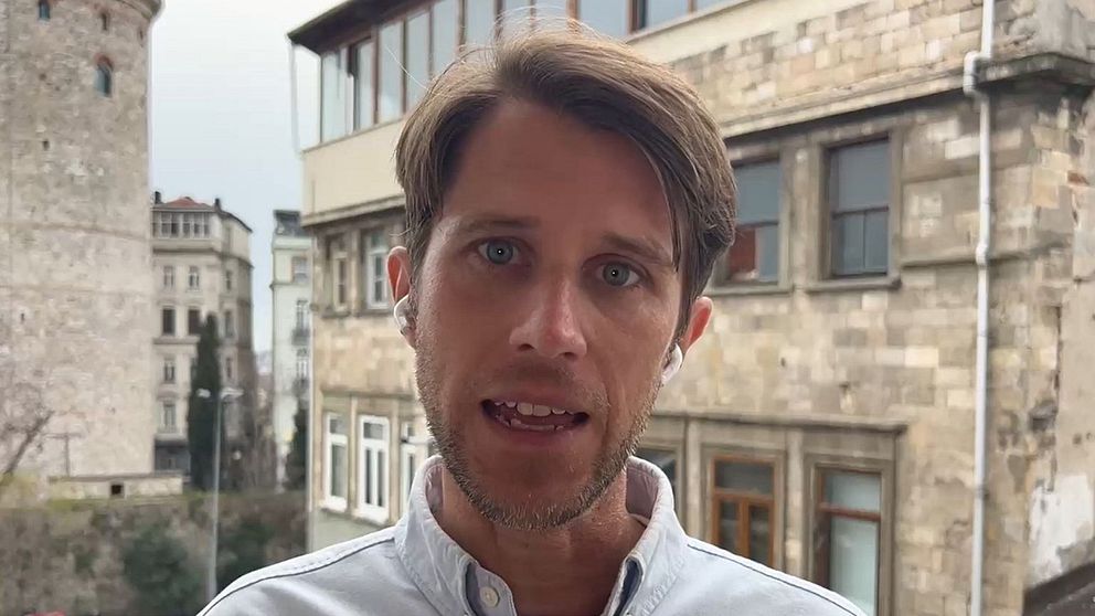 Gammaldags stenbyggnader i stadsmiljö, torn till vänster och träd i bakgrunden.