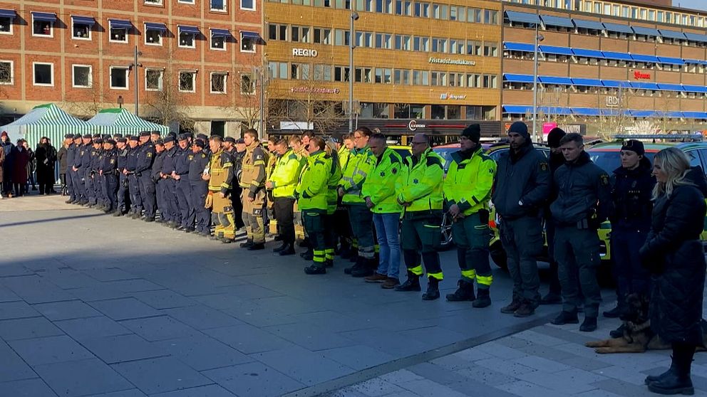 En grupp av räddningspersonal och åskådare står uppställda vid ett torg under en ceremoni eller demonstration framför två större byggnader.
