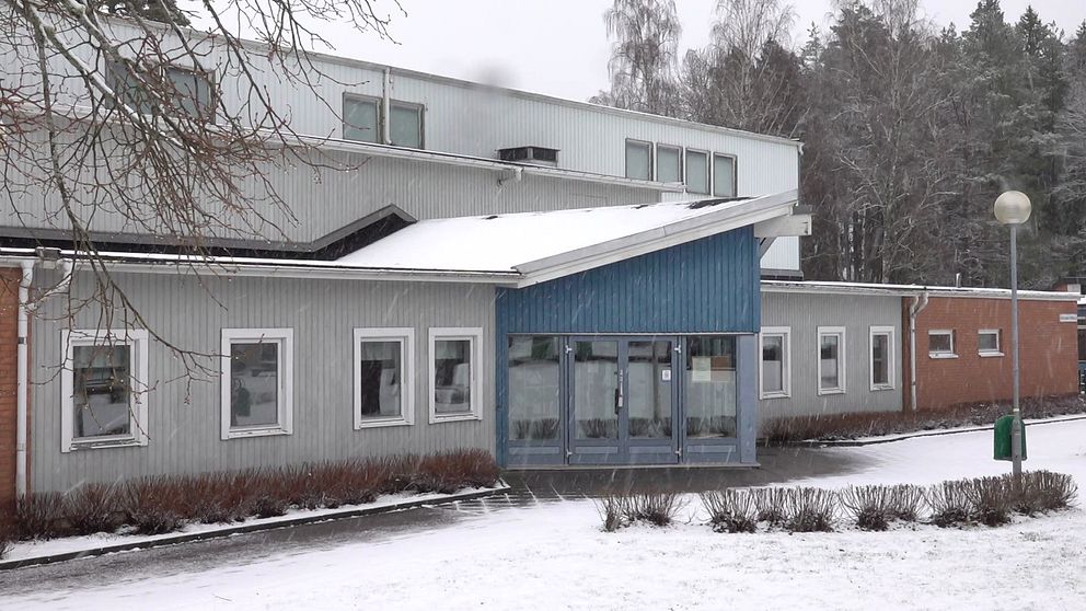 Sandhems skola, en byggnad med gråa väggar och längst bort i bilden är det tegel. På mitten av byggnaden är det blå fasad. På marken ligger det snö.
