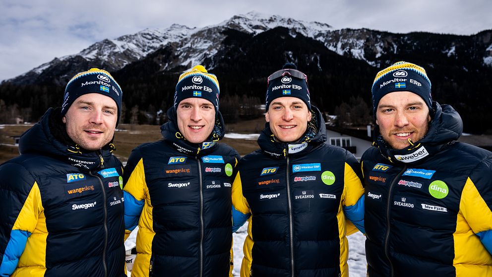Fyra personer i svenska vinterlandslagets jackor står tillsammans med en bergskedja i bakgrunden. De bär varma, matchande jackor med sponsorslogotyper och mössor, omgivna av snötäckta landskap.