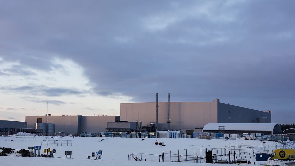 Industriområde med stora byggnader täckta av snö under en molnig himmel. Den främre byggnaden har två rör som sträcker sig mot himlen, medan en mindre byggnad syns till höger. Landskapet präglas av en vintrig miljö och få aktiviteter syns runt området.