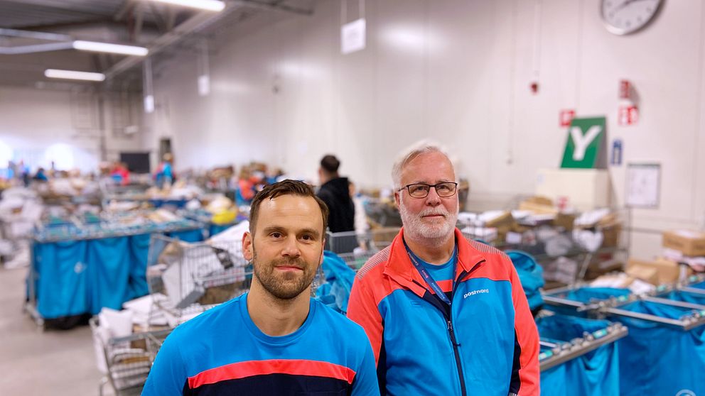 Två personer står i en lagerlokal omgiven av handkorgar och emballage. Den ena personen bär en blå t-shirt med röd detalj, medan den andra har på sig en färgglad jacka. I bakgrunden syns en stor mängd varor och korgar organiserade i rader. En väggklocka hänger på väggen, vilket visar att det är dagsljus.