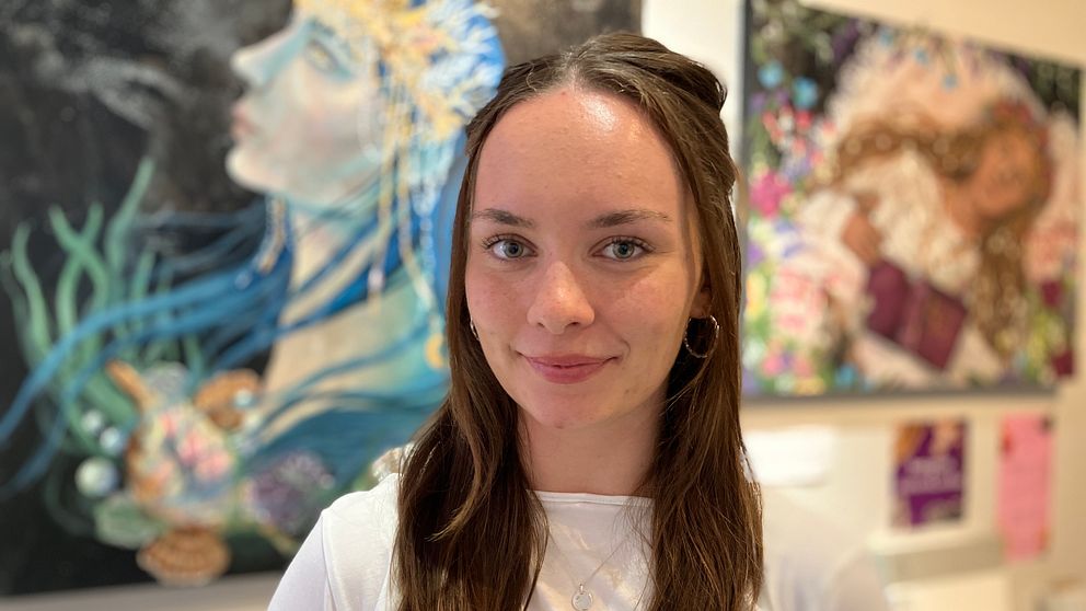 Ung konstnär Evelina Söderström står framför tavlor hon målat.